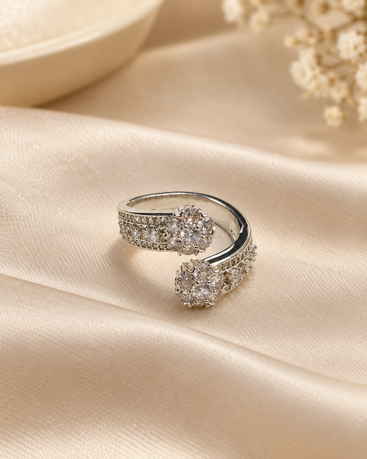 A silver-toned open wrap ring adorned with clear pavé stones and two round cluster settings, displayed on draped champagne satin fabric with soft floral props in the background.